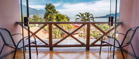 A balcony with a wooden railing, two chairs, and a view of a coastal landscape with mountains and palm trees.