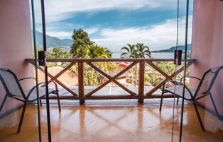 Un balcón con barandilla de madera, dos sillas y vista a un paisaje costero con montañas y palmeras.