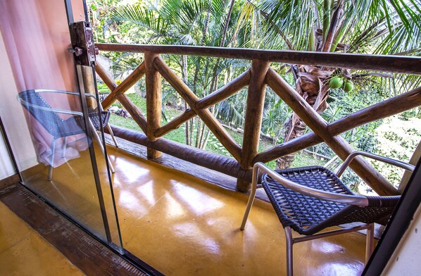Un balcón con puerta de vidrio, una silla de mimbre y vistas a palmeras y follaje.