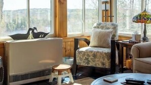 A cozy room with a large window, a radiator, a chair with a cushion, a small stool, and a table with a lamp. - Dragonwood Retreat for Family and Friends (Parkman)