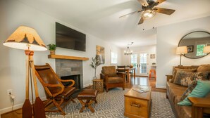 A living room with a fireplace, a ceiling fan, a TV mounted on the wall, and a dining area in the background.