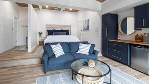 A modern bedroom with a large bed, a blue sofa, a glass coffee table, and a kitchen area with dark cabinets and a refrigerator. - New! Luxurious FLAT lot with private beach, dock, guest suite/apartment. (Lake Ozark)