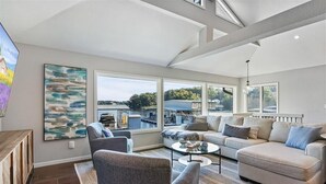 A spacious living room with a large window, a sofa, a coffee table, and a TV. - New! Luxurious FLAT lot with private beach, dock, guest suite/apartment. (Lake Ozark)
