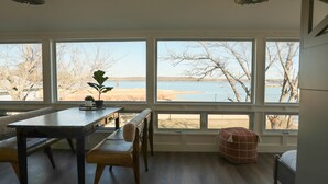 Une pièce dotée de grandes fenêtres donnant sur un plan d’eau, une table en bois avec des chaises et un petit pouf rouge.