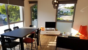 Un espace repas moderne avec une table en bois, des chaises noires et rouges, un meuble blanc avec un téléviseur et de grandes fenêtres donnant sur l’extérieur.