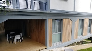 Un bâtiment moderne doté d’un espace extérieur couvert comprenant une terrasse en bois, une table à manger avec des chaises et un petit jardin agrémenté de deux chaises en plastique blanches.