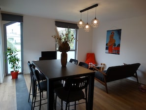 Un espace repas moderne avec une table en bois, des chaises noires et un vase contenant des plantes séchées. On y trouve une fenêtre donnant sur l’extérieur, un tableau accroché au mur et un luminaire suspendu.