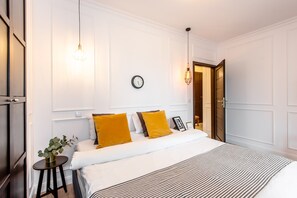 Un dormitorio ordenado con una cama grande, dos mesitas de noche, un reloj de pared y una puerta que da a otra habitación.