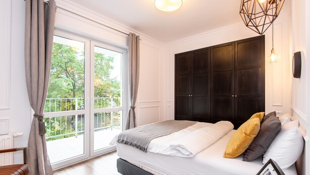 A bedroom with a large window, a bed with pillows, a bedside table, and a hanging light fixture.