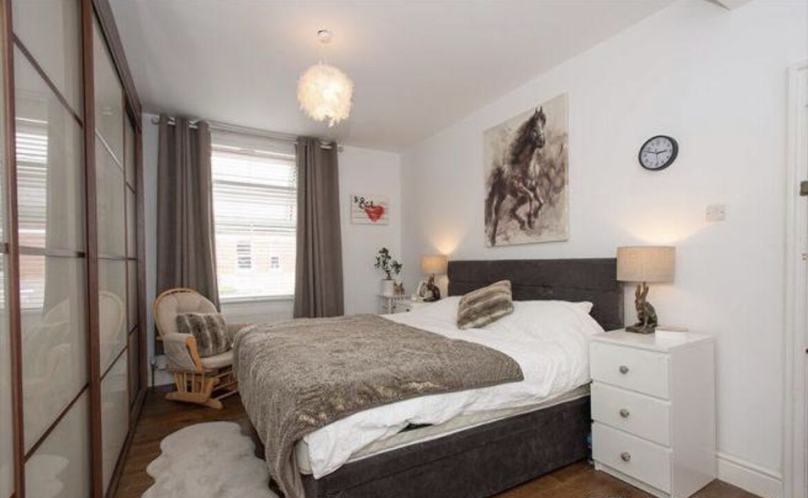 A bedroom with a large bed, a chair, a nightstand, a wall clock, and a window with curtains.