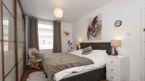 A bedroom with a large bed, a chair, a nightstand, a wall clock, and a window with curtains.