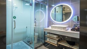 Une salle de bain moderne dotée d’une douche à parois de verre, d’un meuble-lavabo avec un plan de travail en marbre et d’un grand miroir surmonté d’un luminaire circulaire.