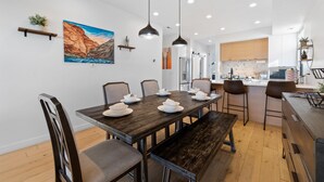 A modern dining area with a wooden table set for six, surrounded by chairs. A bench is placed at the end of the table. The room features a white ceiling with recessed lighting and a large painting of a landscape on the wall.