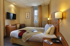 A hotel room with a bed, bedside table, lamp, TV, and a window with curtains.