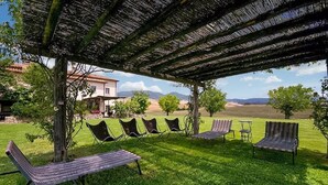 A covered outdoor seating area with multiple lounge chairs and a table, set against a backdrop of a house and a grassy field.