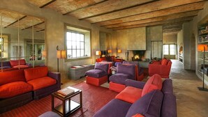 A spacious living room with high ceilings and exposed brick walls, featuring a fireplace, multiple seating areas, and a large window providing natural light.