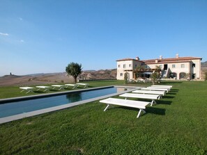 Una piscina con sillones de descanso, un edificio con tejado de tejas rojas y un área verde con colinas al fondo.