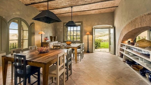 A rustic dining area with a wooden table and chairs, a black pendant light, and a view of the outdoors through a large window.