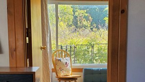 A room with a wooden desk, a chair with a floral cushion, a blue sofa, and a view of greenery through a large window. - Tip of the Peninsula Lake House Retreat (South Bend)
