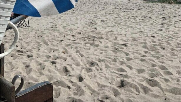 Ein Strand mit einem weißen und blauen Streifen-Strandsonnenschirm, einer Holzbank und einer felsigen Küstenlinie unter einem bewölkten Himmel.