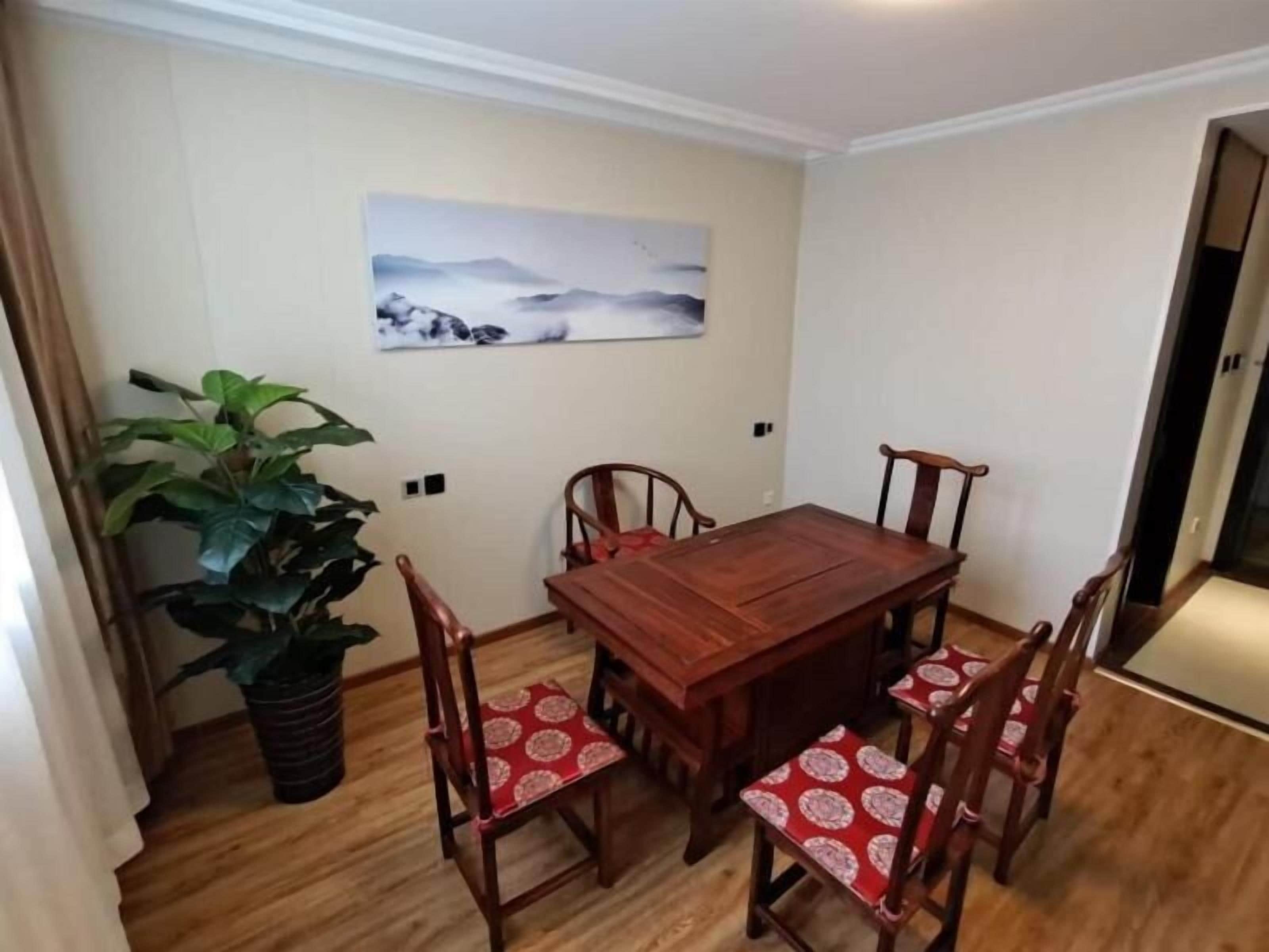 A dining area with a wooden table and four chairs, a large potted plant, and a wall-mounted painting.