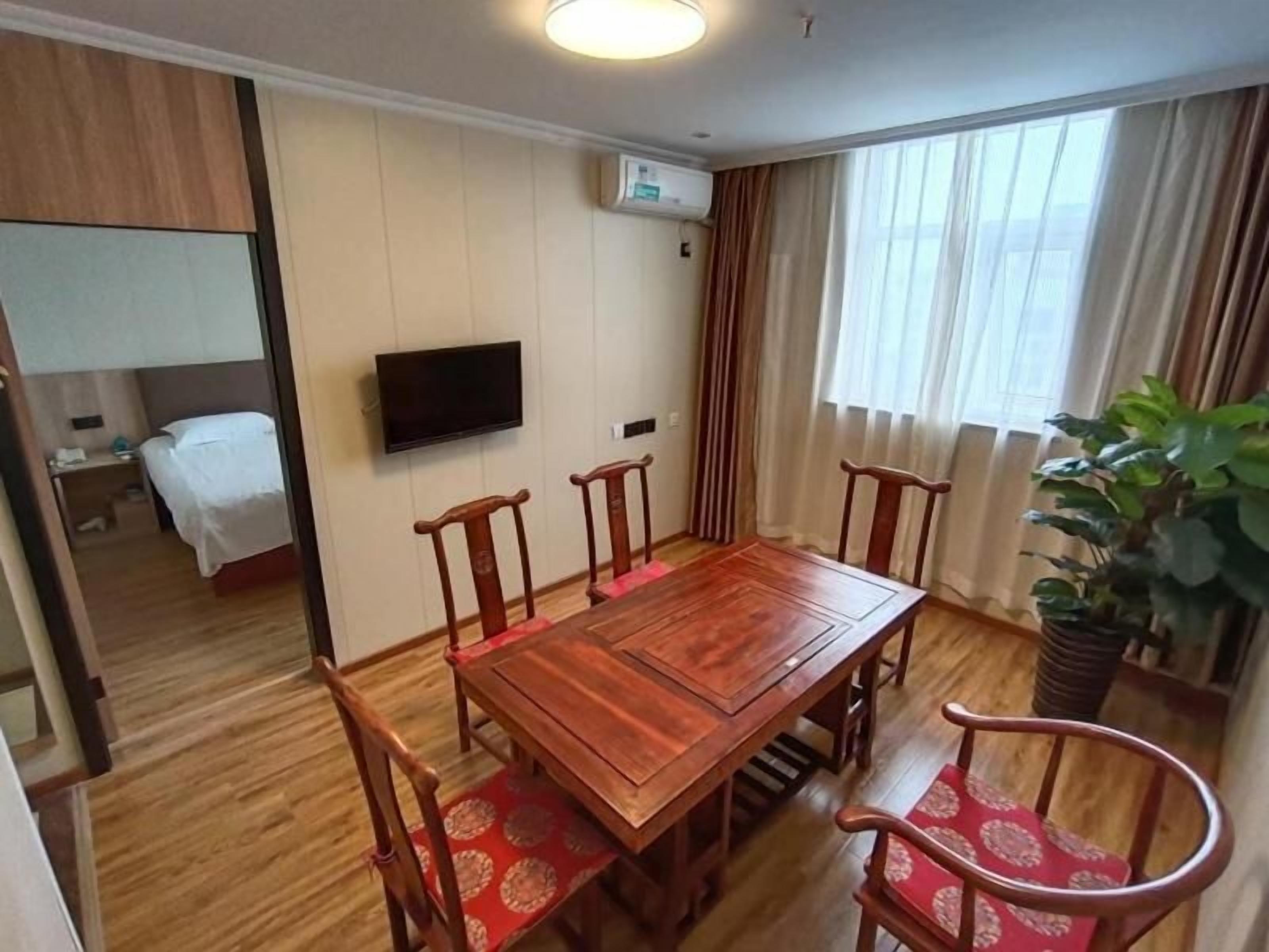 A hotel room with a wooden dining table, four chairs, a TV mounted on the wall, a bed, and a potted plant.
