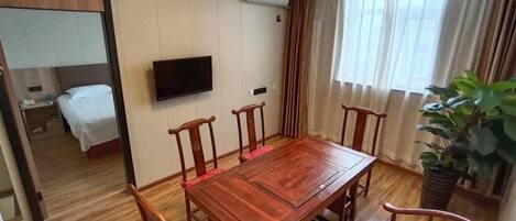A hotel room with a wooden dining table, four chairs, a TV mounted on the wall, a bed, and a potted plant.