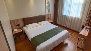 Une chambre d’hôtel avec un grand lit, une tête de lit en bois, une table de chevet avec un téléphone, un plaid vert et une fenêtre avec des rideaux.