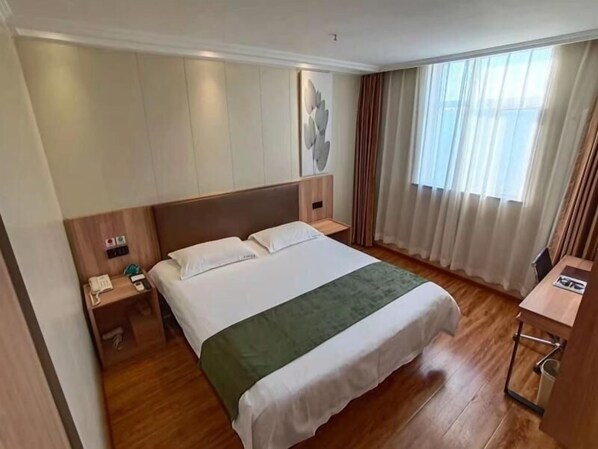 Une chambre d’hôtel avec un grand lit, une tête de lit en bois, une table de chevet avec un téléphone, un plaid vert et une fenêtre avec des rideaux.