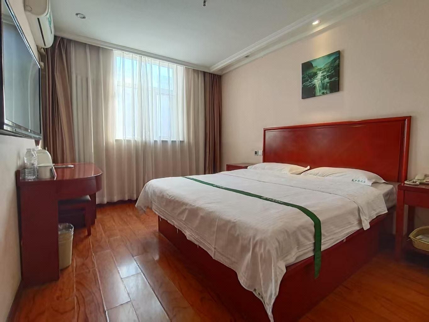 A hotel room with a wooden floor, a bed with white bedding, a red headboard, a bedside table, a nightstand, a painting on the wall, and a window with curtains.