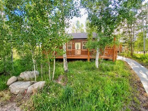 Une cabane dotée d’une terrasse en bois et d’un porche, entourée d’arbres et d’une zone herbeuse.