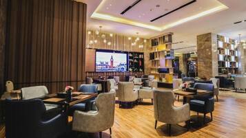 A modern hotel lobby with a bar area, seating arrangements, and a television screen displaying a cityscape.