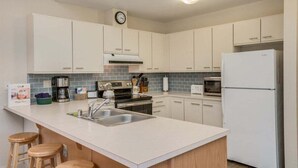 Une cuisine moderne avec des meubles blancs, un îlot central doté de tabourets, un réfrigérateur et une horloge murale.
