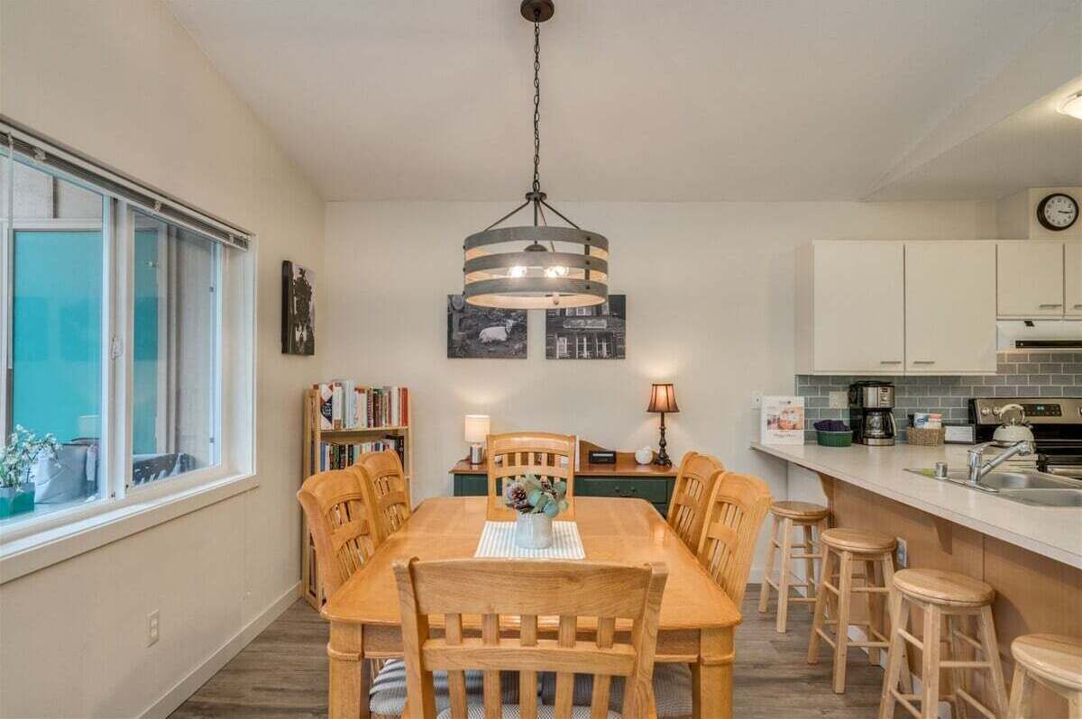 Un comedor con mesa y sillas de madera, una lámpara colgante, una estantería y una zona de cocina con armarios y un fregadero.