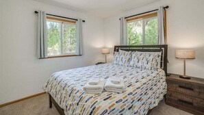 Ett sovrum med en säng, två sänglampor, en byrå i trä och två fönster med gardiner.