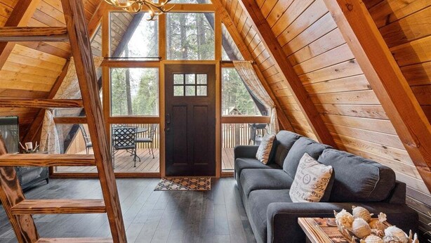 A cozy cabin with a loft bedroom, a dark wooden door, a gray sofa with decorative pillows, and a wooden staircase. - Pet-friendly A-Frame Near Plain & Leavenworth (Leavenworth)