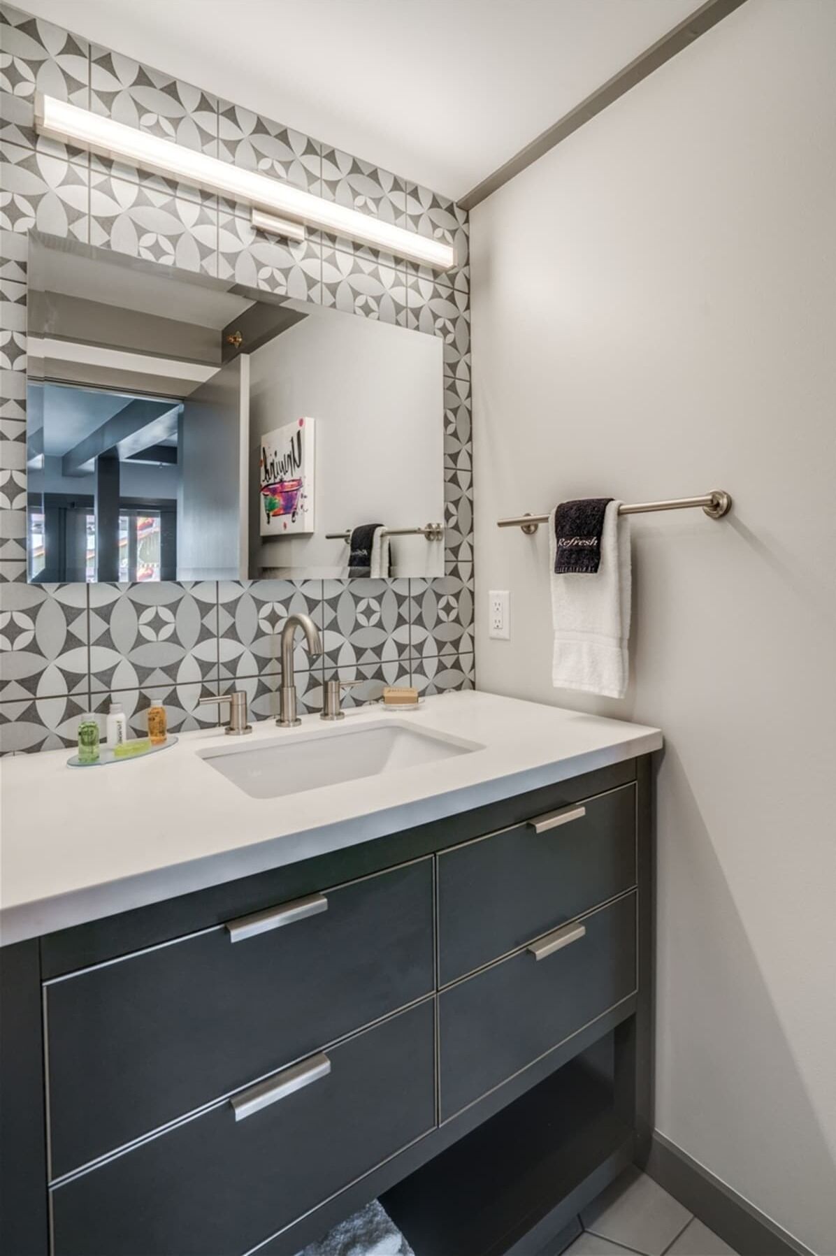 A modern bathroom with a geometric patterned tile backsplash, a large mirror, a sink with a faucet, and a towel rack.