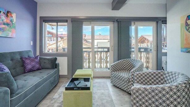 A living room with a grey sofa, patterned armchairs, a small table, and a sliding glass door leading to a balcony with a view of buildings. - Downtown Leavenworth King Studio - Parking + Balcony (Leavenworth)