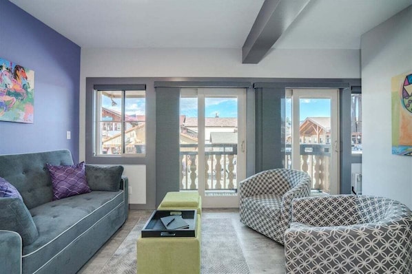 En stue med en grå sofa, mønstrede lænestole, et lille bord og en skydedør, der fører ud til en balkon med udsigt til bygninger.