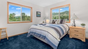 Ein Schlafzimmer mit einem großen Bett, zwei Nachttischen, einer Stehlampe, einem kleinen Hocker und Blick auf eine grüne Landschaft durch die Fenster.