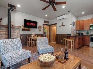 A living room with patterned sofas, a wooden coffee table with snacks, and a kitchen area with wooden cabinets.