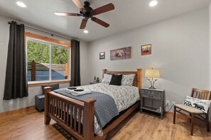 Un dormitorio con una cama de madera, un ventilador de techo, una mesita de noche con lámpara y una ventana con cortinas.