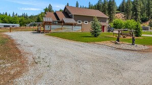 A building with a sloped roof, a gravel driveway, and a fenced area with two sculptures. - Cozy Mountain Retreat Near Leavenworth w/ Private Patio + BBQ (Leavenworth)