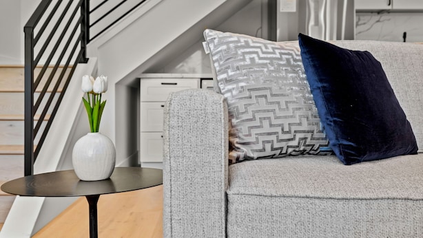 En moderne stue med en grå sofa, en pudde med et mønster, en vase med hvide blomster og en trappe i sort metal.