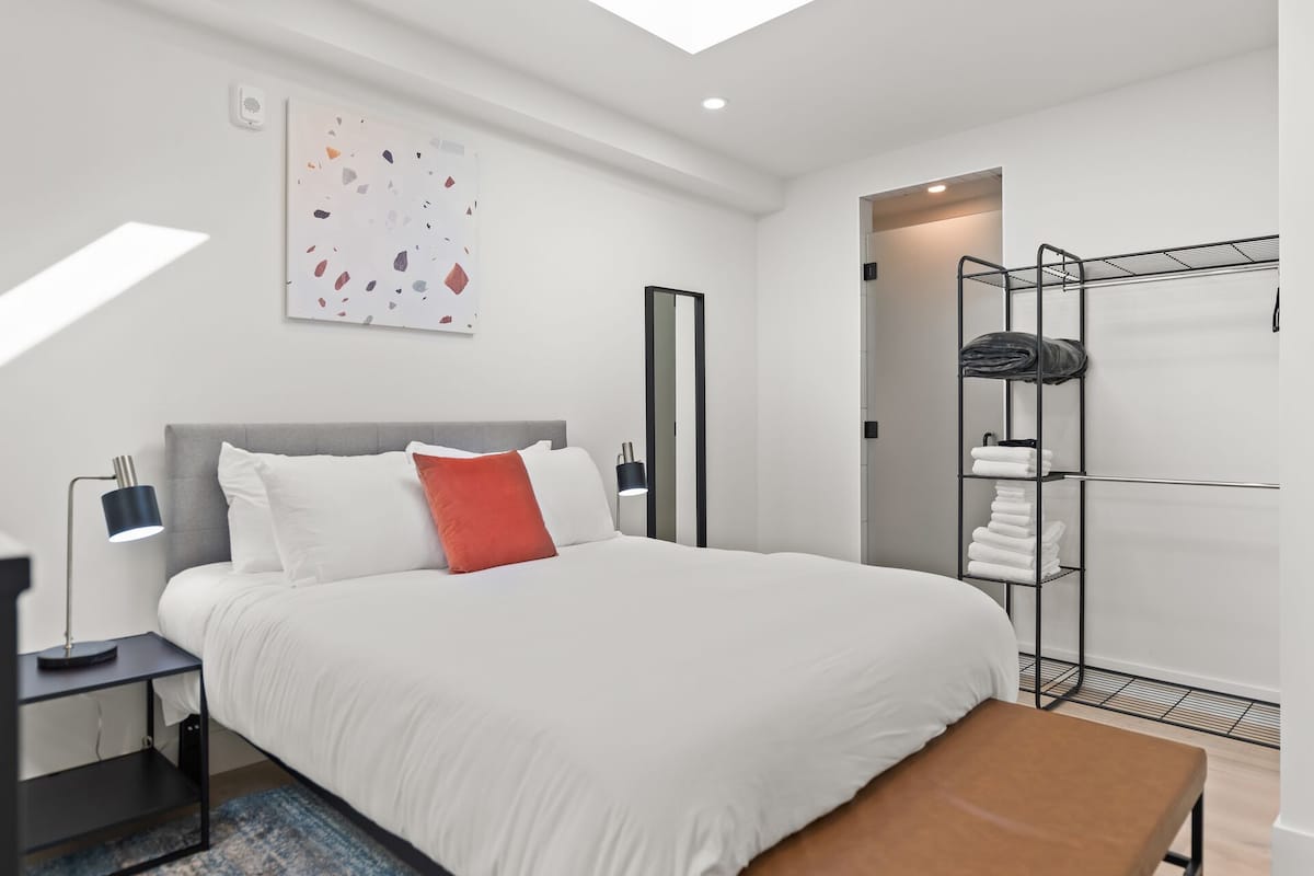 Moderni hotellihuone, jossa on suuri sänky, yöpöytä lampulla, penkki, vaatekaappi, jossa on henkarit, ja kattovalaisin.
