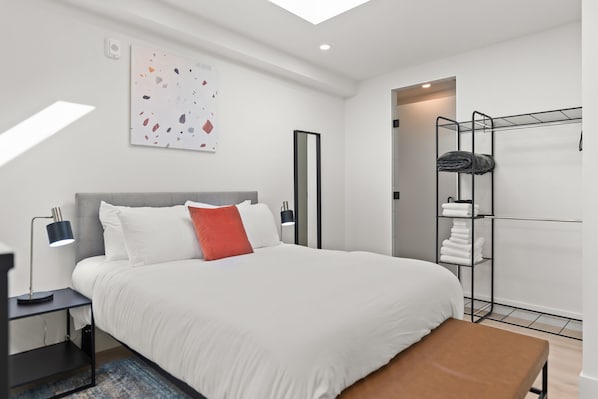 Habitación de hotel moderna con una cama grande, una mesita de noche con lámpara, un banco, un armario con ropa colgada y una luz en el techo.