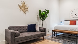 Et moderne soveværelse med en grå sofa, en træbænk, en stor grøn plante og en seng med hvide sengetæpper og orange puder.