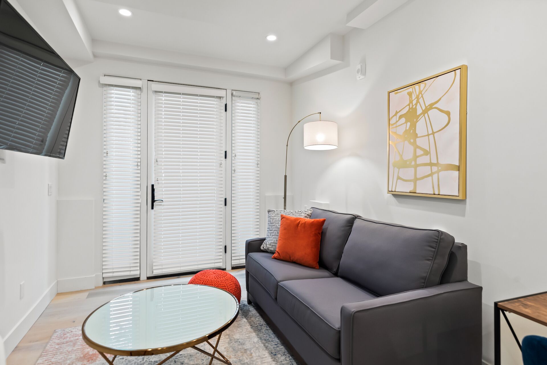 Una sala de estar moderna con un sofá gris, una mesa de centro de vidrio y un taburete rojo. Hay un ventanal con persianas, una lámpara de pie y una obra de arte abstracta en la pared.
