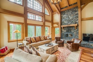 A spacious living room with a stone fireplace, large windows, and wooden beams. - Norge Haus - Riverfront Extraordinary Home (Leavenworth)