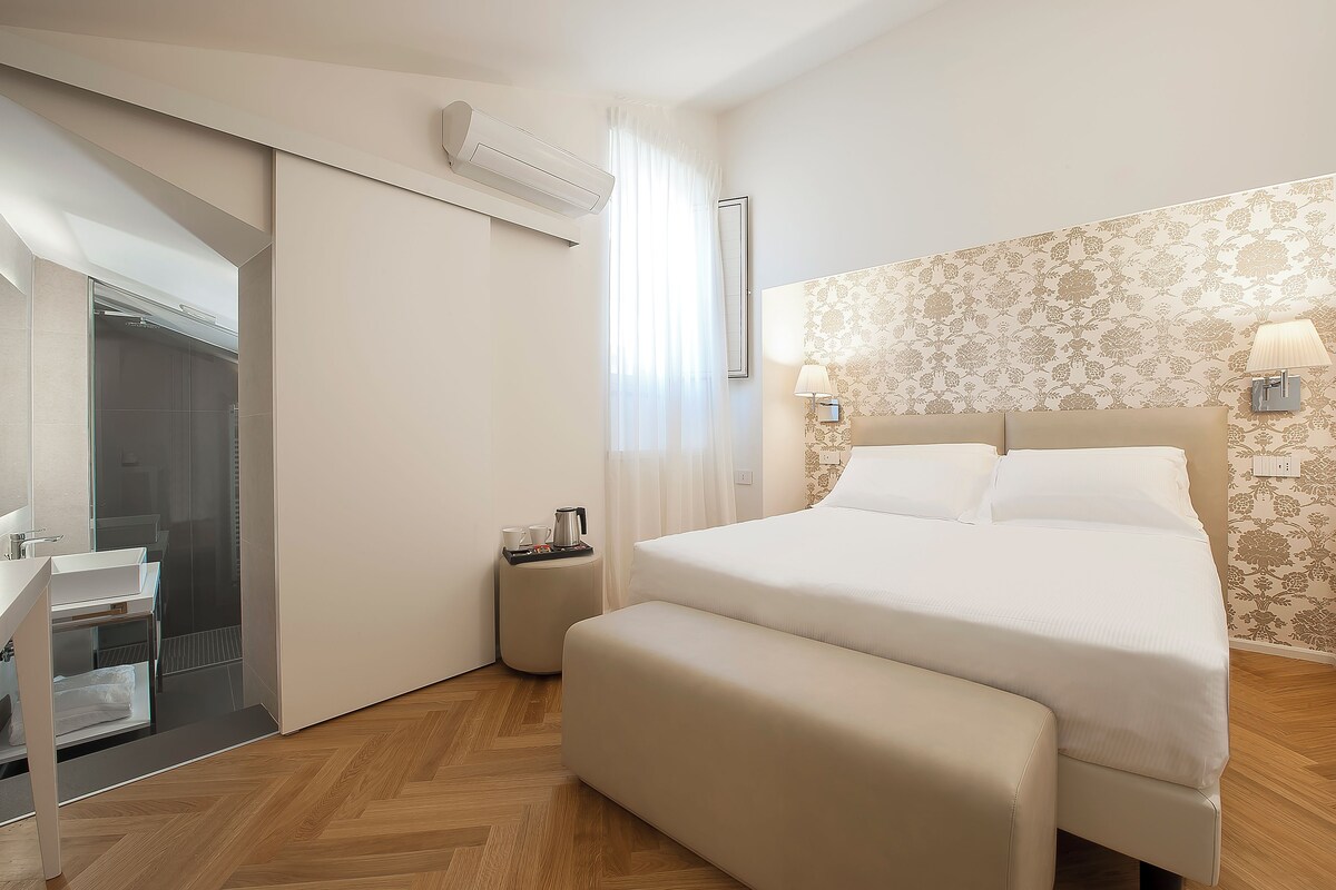 A modern hotel room with a large bed, a bedside table with a lamp, a small bedside table with a cup and a book, and a sliding door leading to a shower area.